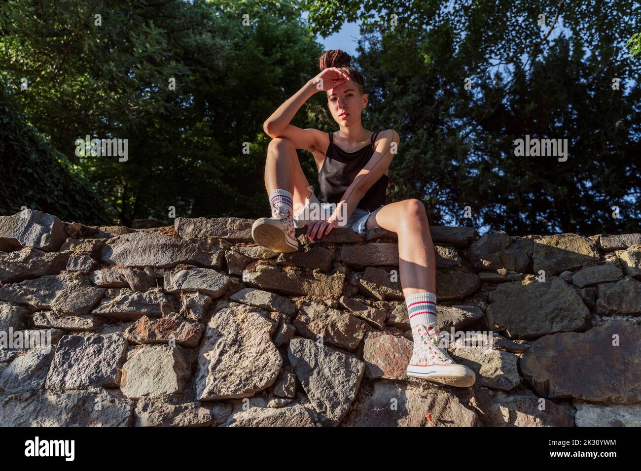 Hipster non binario seduto su un muro di pietra Foto Stock