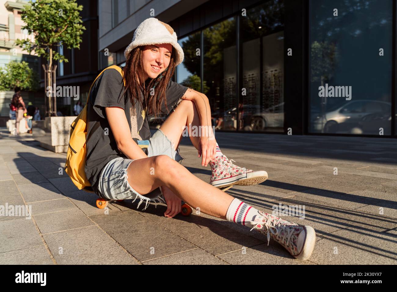 Sorridente persona non binaria seduta sullo skateboard sul sentiero Foto Stock