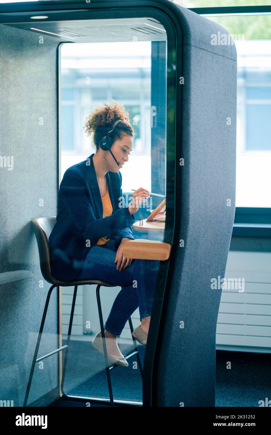 Donna d'affari matura con cuffie che utilizzano un tablet PC seduto in una cabina insonorizzata sul posto di lavoro Foto Stock