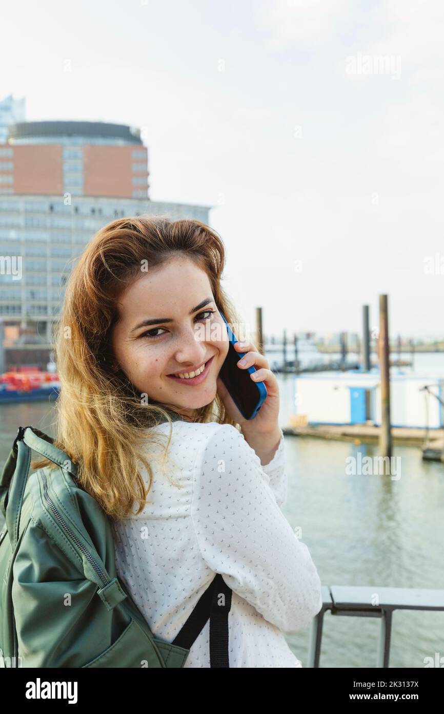 Donna sorridente che parla su smartphone in piedi in ringhiera al porto di Amburgo, Germania Foto Stock