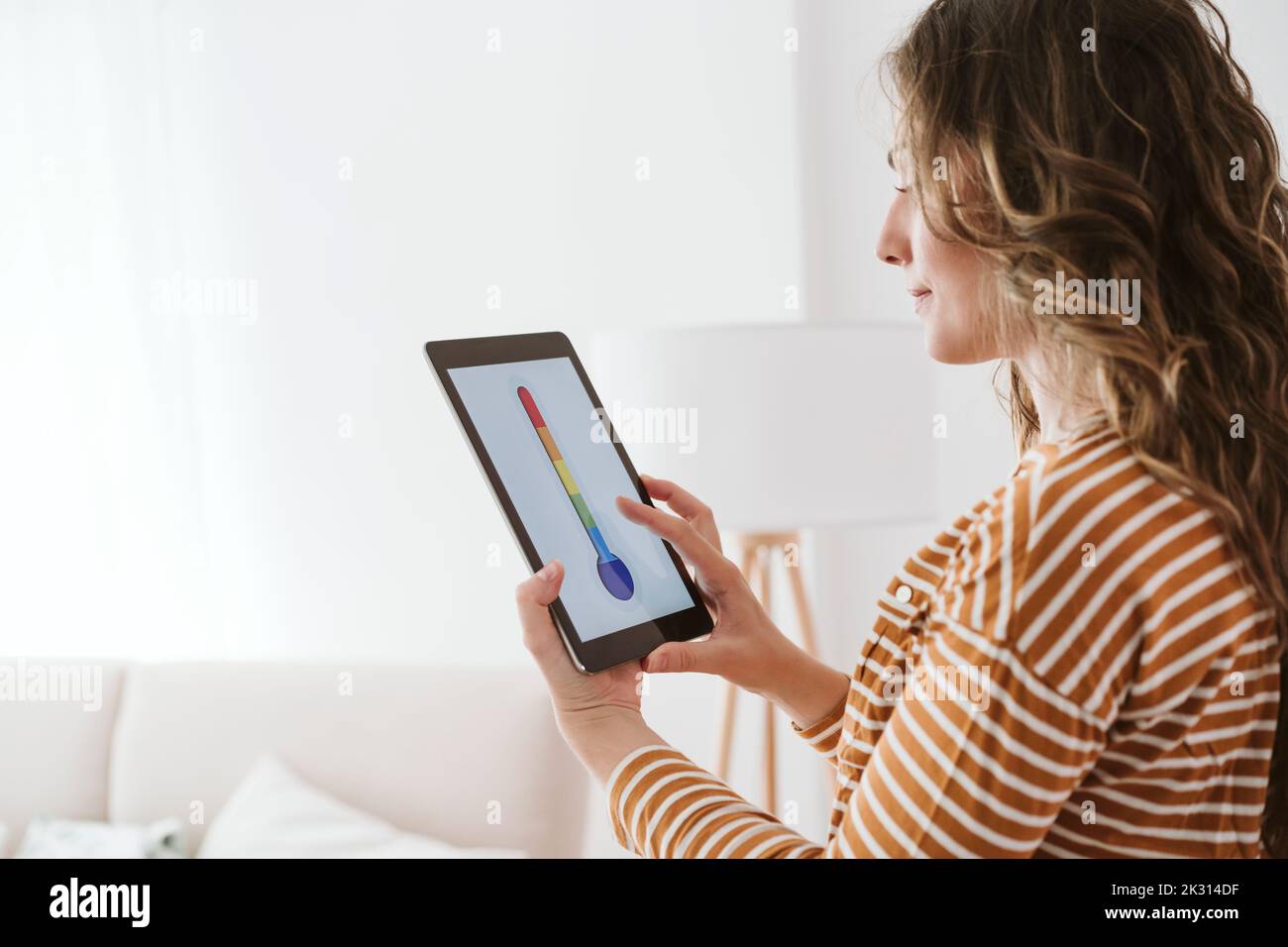 Giovane donna che usa un tablet digitale con l'icona del termometro nel salotto Foto Stock