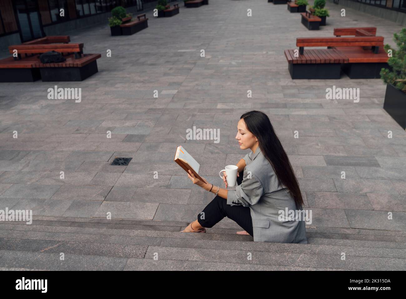 Donna d'affari con tazza di caffè libro di lettura su gradini al parco ufficio Foto Stock