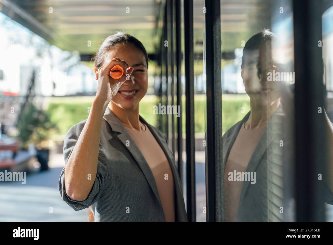 Donna d'affari sorridente che guarda attraverso carta arrotolata da riflessione su parete di vetro Foto Stock