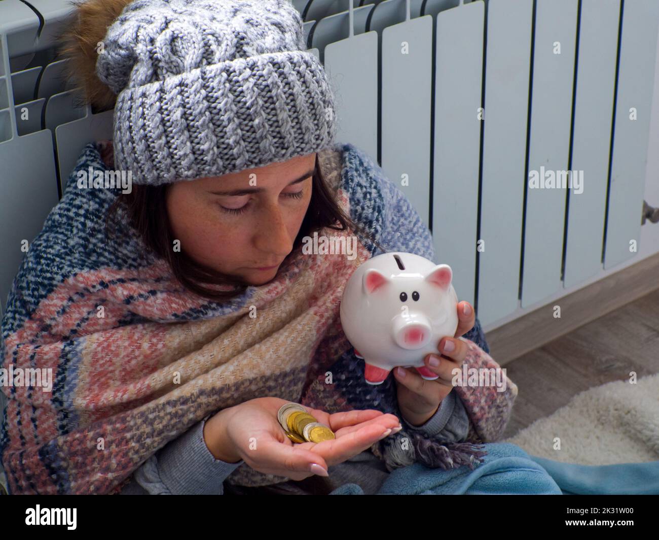 Donna che conta monete in euro e tiene il piggy bank vicino alla batteria a casa. Donna vestita calorosamente è sconvolta circa il prezzo elevato di riscaldamento. Costoso it Foto Stock