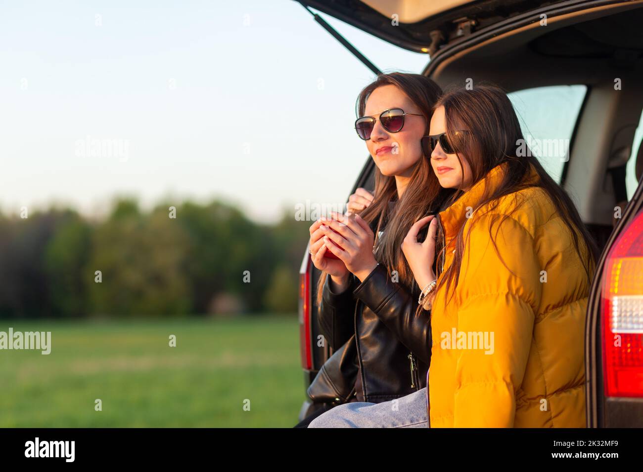 Madre e figlia si accampano su una collina e ammirano il tramonto mentre si siedono nel bagagliaio dell'auto Foto Stock