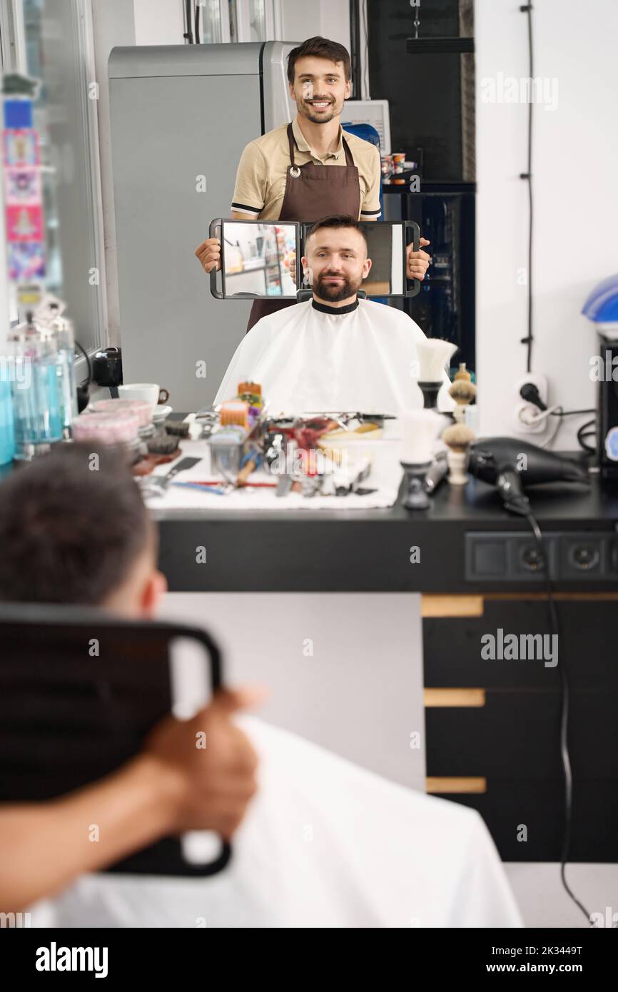 Barbiere soddisfatto che presenta i risultati del taglio dei capelli ad un cliente soddisfatto Foto Stock