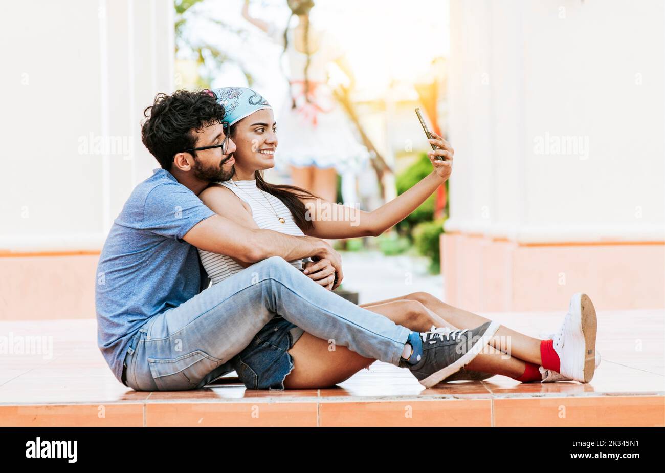 Giovane coppia che prende un selfie seduto sul pavimento. Giovane coppia latina innamorata seduta sul pavimento prendendo un selfie all'aperto. Concetto di coppia felice Foto Stock