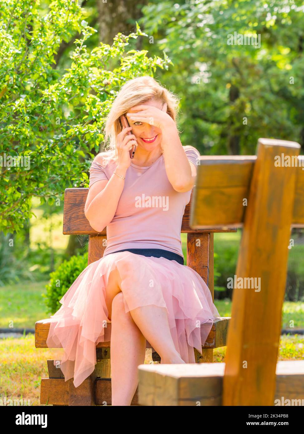 Donna bionda seduta nel parco che sbirciava davanti alla fotocamera sorridendo mentre parla utilizzando il telefono smartphone Foto Stock