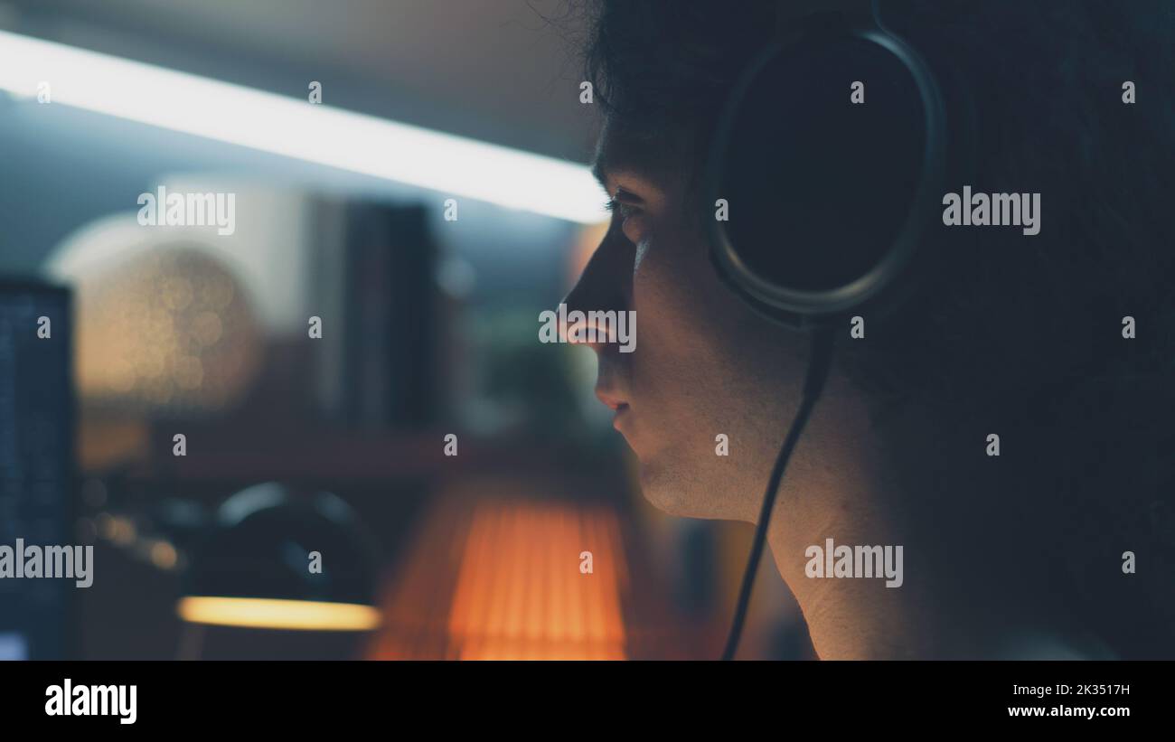 Uomo in cuffia che naviga su Internet e sui social network sul computer di casa di notte e ascolta musica o lavora online da freelance Foto Stock