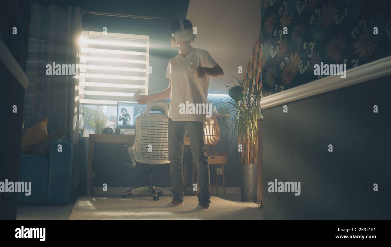 Un ragazzo che si muove mentre gioca a un videogioco a casa utilizzando il visore VR e i controller wireless nel tempo libero. Dispositivo per realtà virtuale. Cyberspazio e metaverse Foto Stock