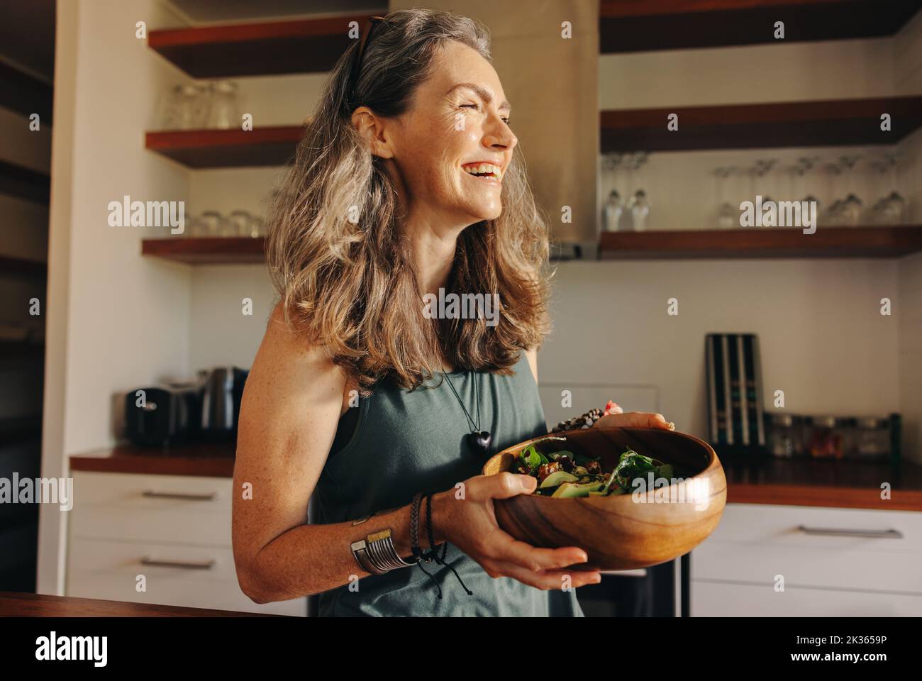 Donna anziana sorridente felice mentre tiene una ciotola di buddha nella sua cucina. Donna anziana felice che si serve un pasto vegano sano a casa. Donna matura Foto Stock