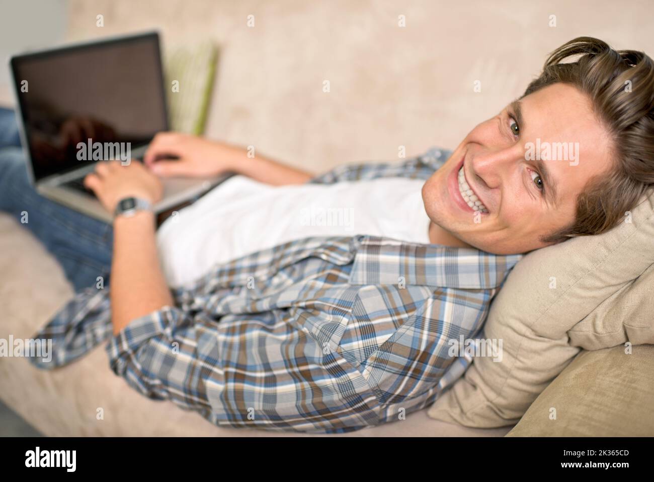 Troppo pigro per alzarsi. Un bel giovane che usa il suo computer portatile mentre si sdraiava su un divano. Foto Stock