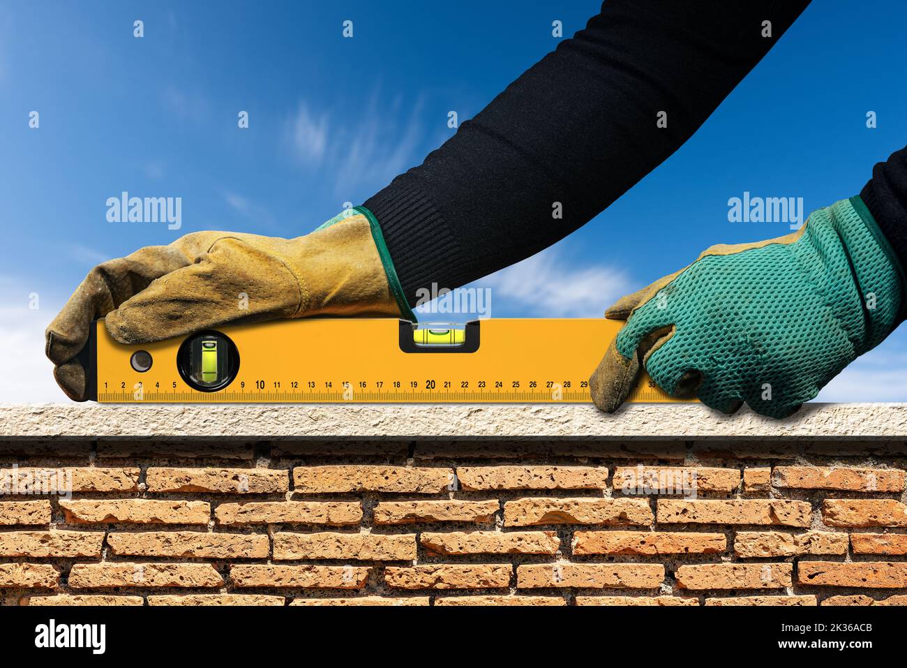 Operatore manuale con guanti di lavoro protettivi, che tiene una livella a bolla d'aria arancione, sopra una parete in mattoni e marmo con cielo blu sullo sfondo. Foto Stock