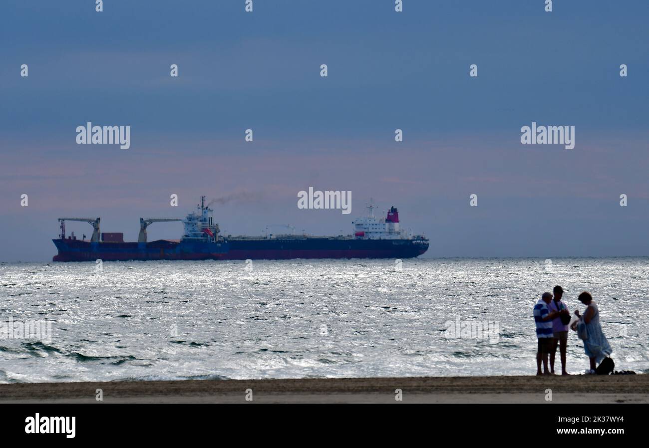Tre bagnanti chiacchierano sulla spiaggia al tramonto con una grande nave portacontainer all'orizzonte. Foto Stock