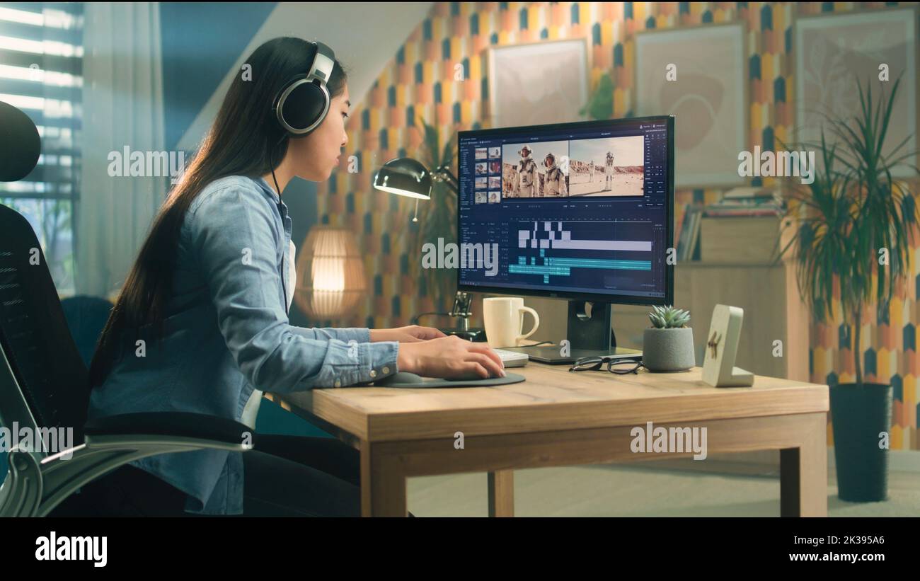 Donna asiatica in cuffie editing video per blog sullo spazio in software professionale sul computer, mentre seduto al tavolo a casa e lavorando in remoto Foto Stock