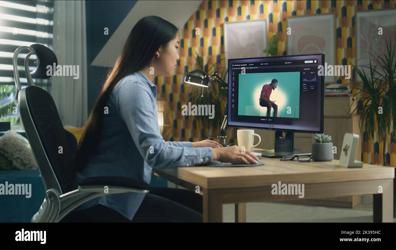 Donna asiatica che modifica foto nel photoshop per i clienti sul personal computer mentre si siede al tavolo in ufficio e lavora in remoto Foto Stock