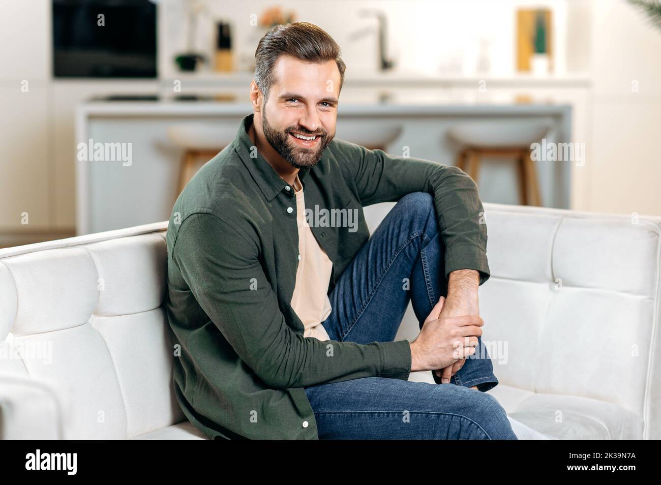 Foto di un bell'elegante uomo caucasico bearded, in abiti casual, seduto su un divano in un accogliente soggiorno, rilassarsi dopo una giornata di lavoro, guardando la macchina fotografica, sorridendo felicemente Foto Stock