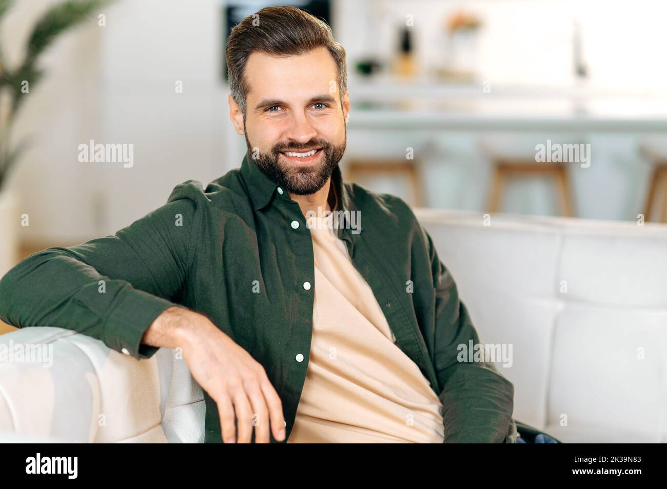 Ritratto di un bell'attraente uomo caucasico moderno bearded, in abiti casual elegante, siede su un divano nel soggiorno, guarda la macchina fotografica, sorrisi amichevole Foto Stock