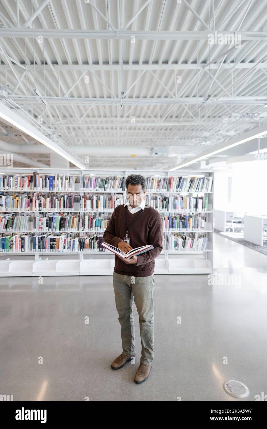 Professore universitario maschile con libro di testo in biblioteca Foto Stock
