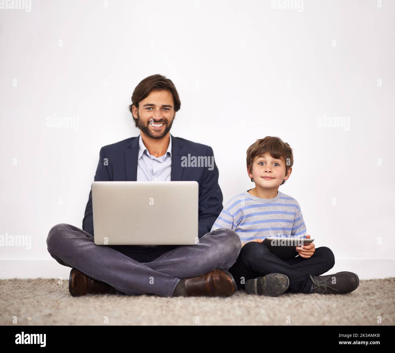 Aumentare la generazione digitale. Un padre e un figlio seduti a terra contro una parete con un notebook e un tablet digitale. Foto Stock