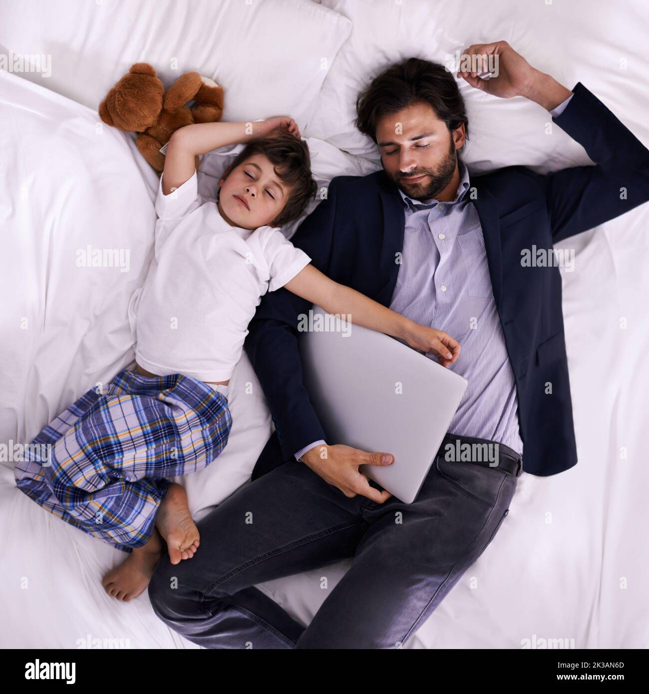 Essere un solo padre può essere estenuante. Un padre e un figlio dormono su un letto mentre suo padre è ancora completamente vestito e tiene un computer portatile. Foto Stock