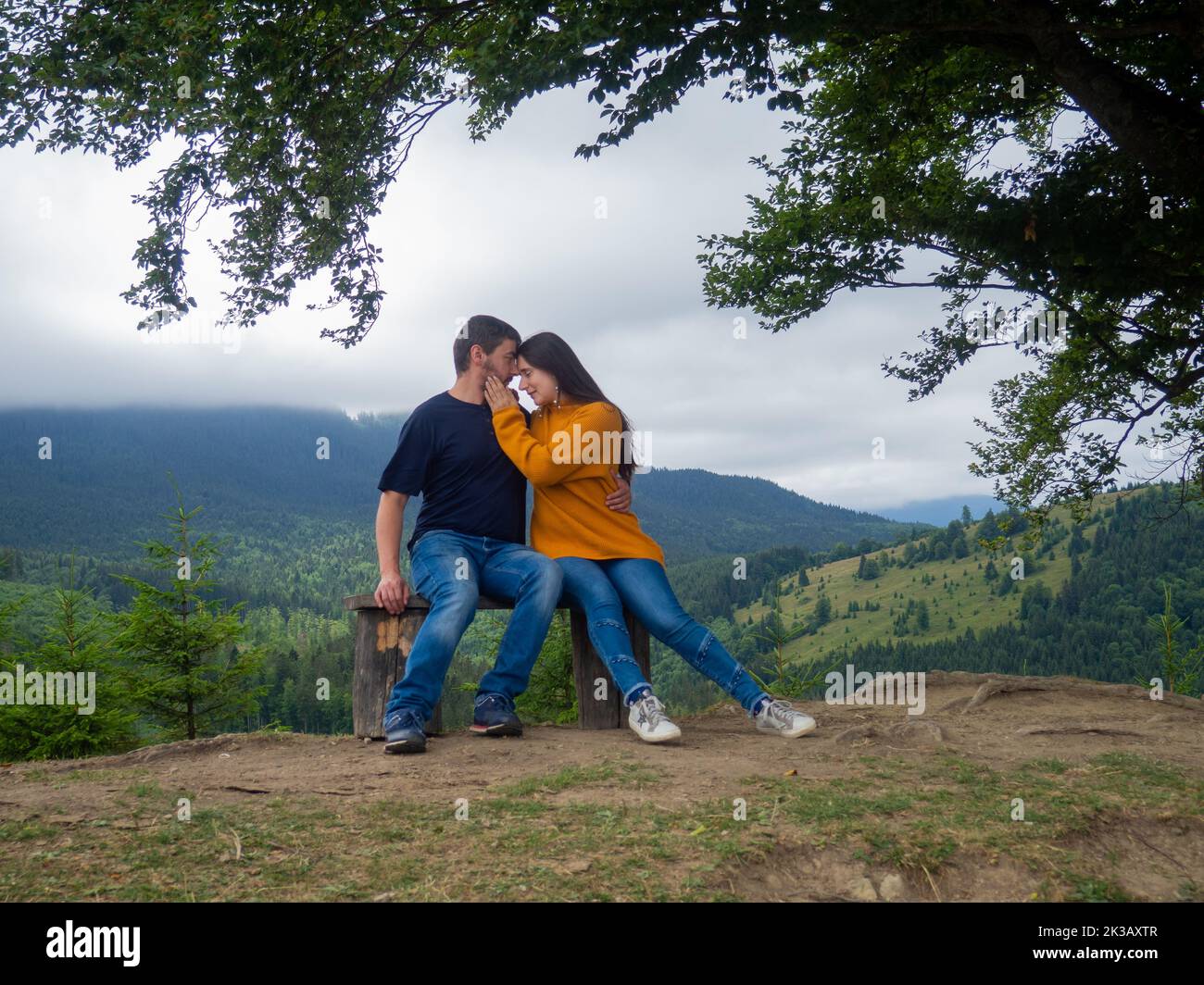 Giovane uomo e donna che ammirano una vista mozzafiato mentre si siede sulla panchina in montagna. Bella coppia in abiti casual sedersi sotto un grande albero con la parte anteriore Foto Stock