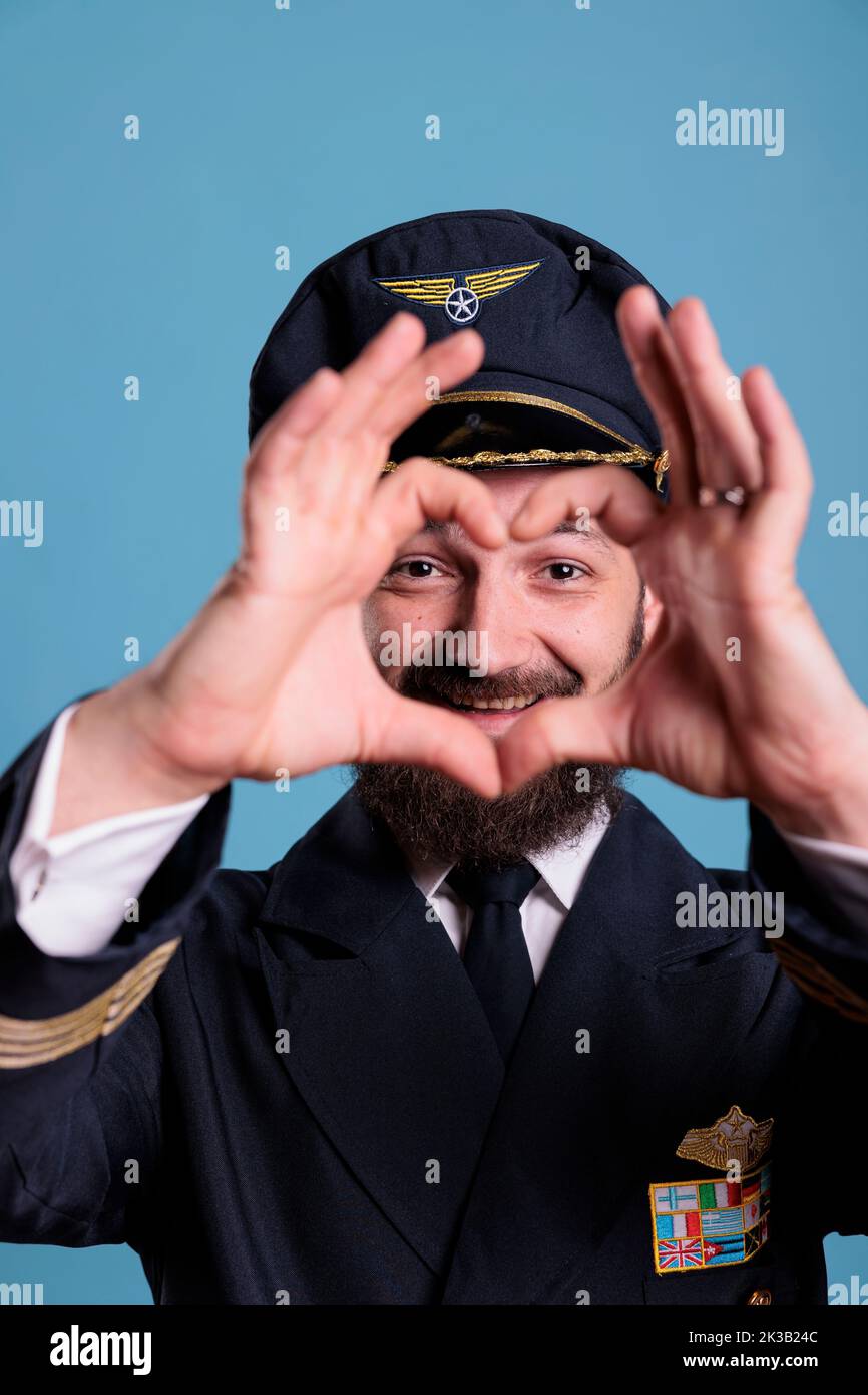 Pilota sorridente che mostra simbolo d'amore a forma di cuore con le dita, gesto romantico concettuale. Amichevole aviatore di aeroplano in uniforme esprimente sentimenti di affetto vista frontale scatto medio Foto Stock