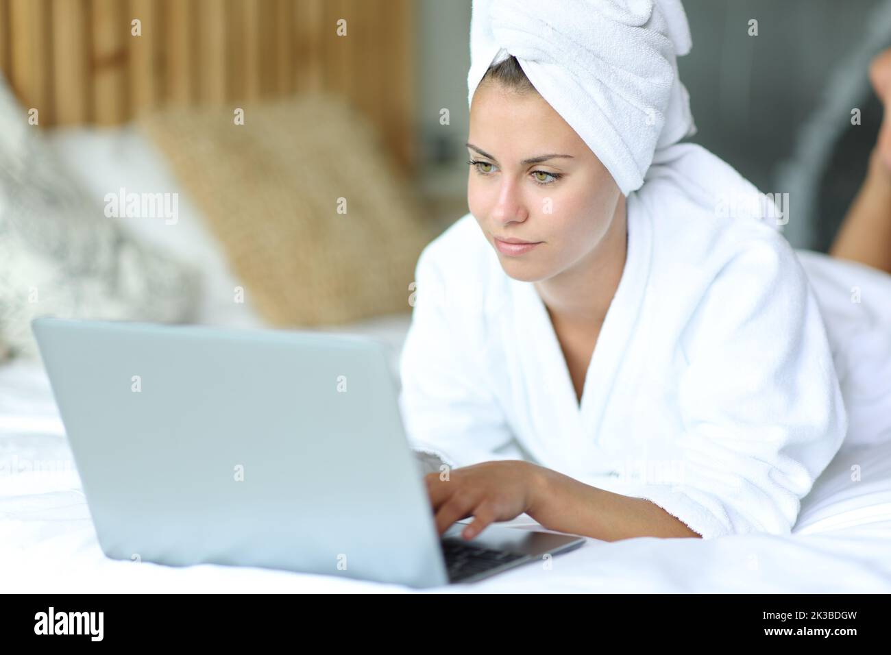 Bella donna sdraiata su un letto dopo aver fatto la doccia usando il notebook a casa Foto Stock