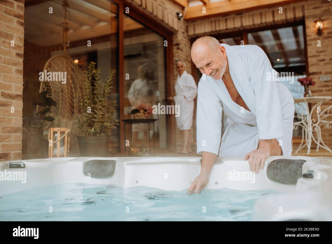 Un uomo anziano che controlla la temperatura dell'accappatoio nella vasca idromassaggio all'aperto, si prepara a fare il bagno con sua moglie. Foto Stock