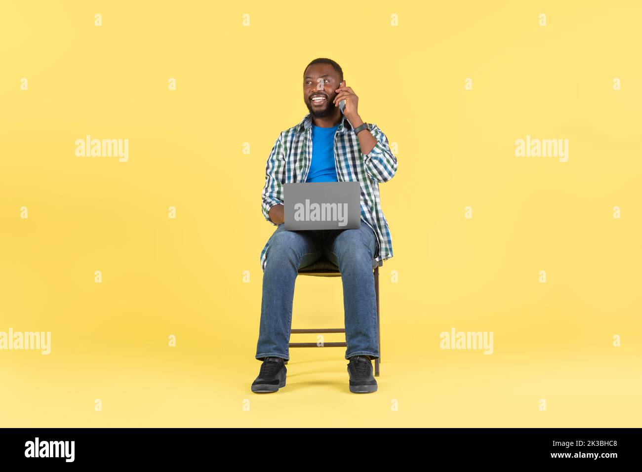 African American Guy che usa laptop parlando su cellulare, sfondo giallo Foto Stock