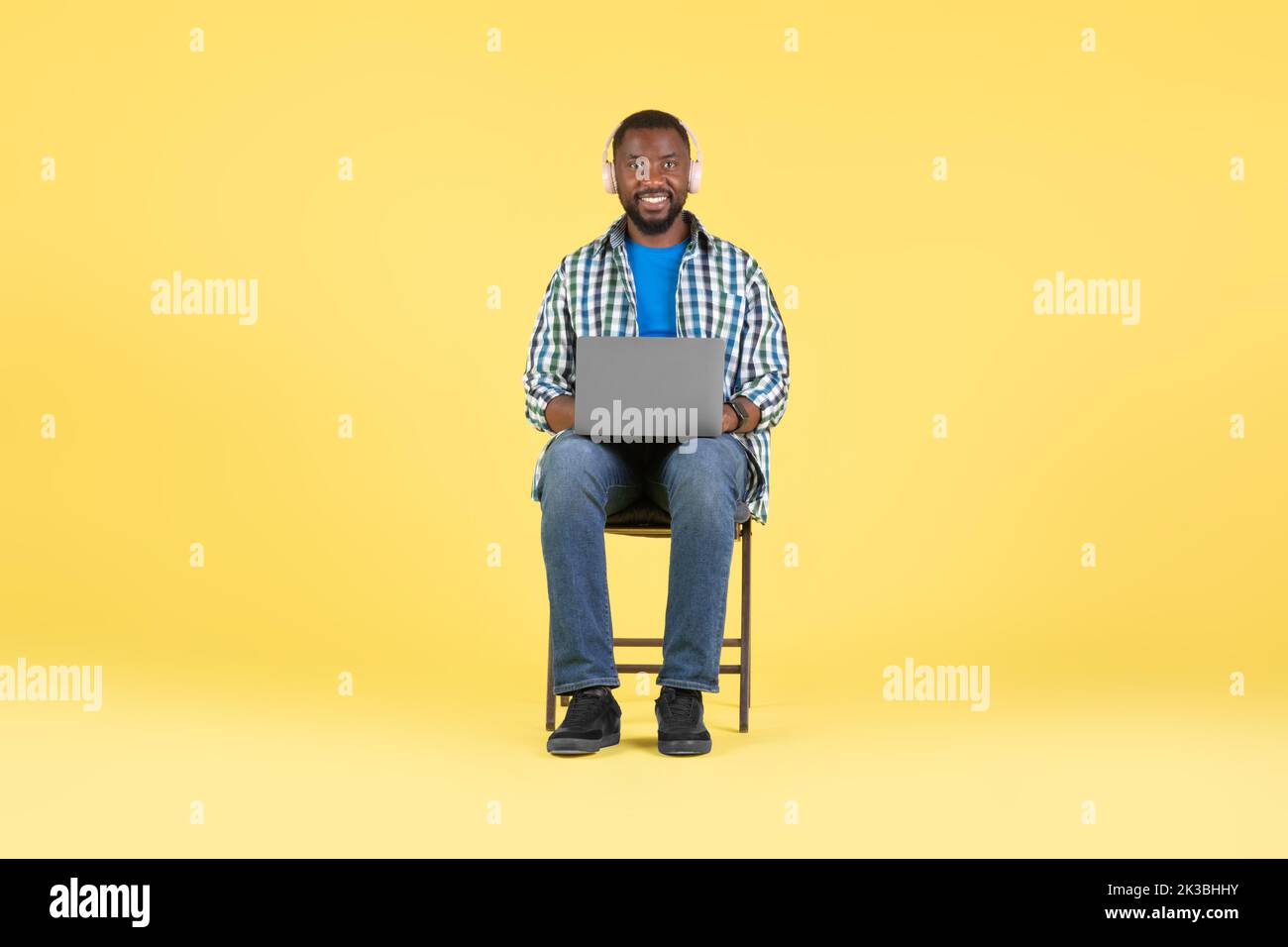 Uomo africano che usa il laptop che indossa gli auricolari seduti su sfondo giallo Foto Stock