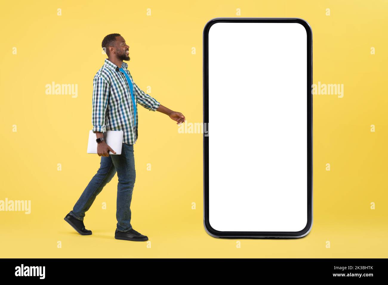 African Man Holding laptop Walking vicino a Big Cellphone, sfondo giallo Foto Stock