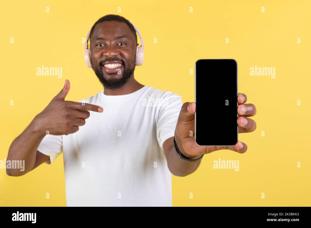 Afro americano maschio che mostra lo schermo del telefono che indossa gli auricolari, sfondo giallo Foto Stock
