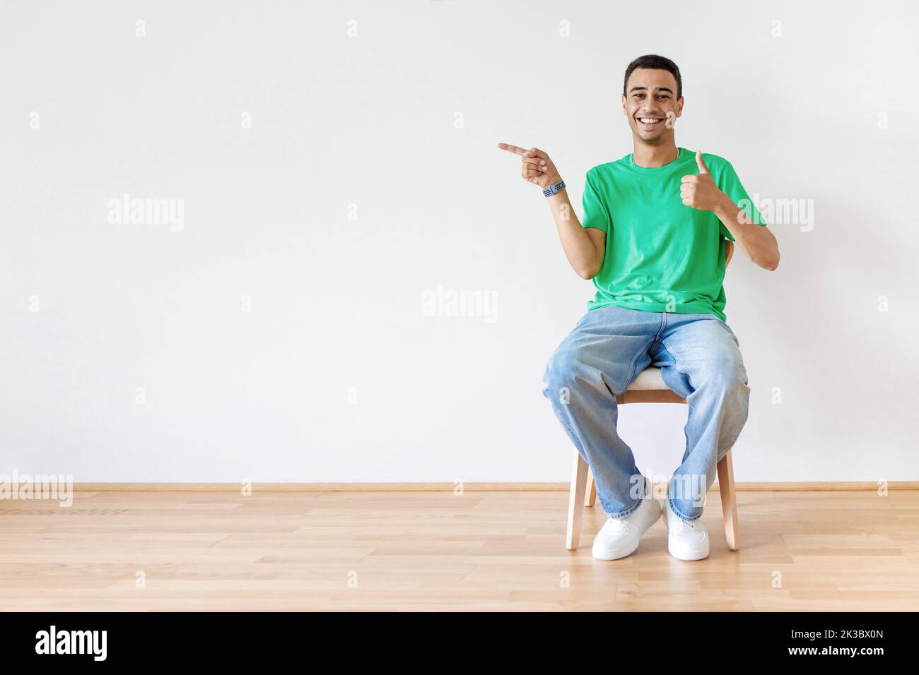 Felice uomo arabo gesturing pollice su e puntando il dito da parte a spazio libero, seduta su sedia su sfondo parete chiaro Foto Stock