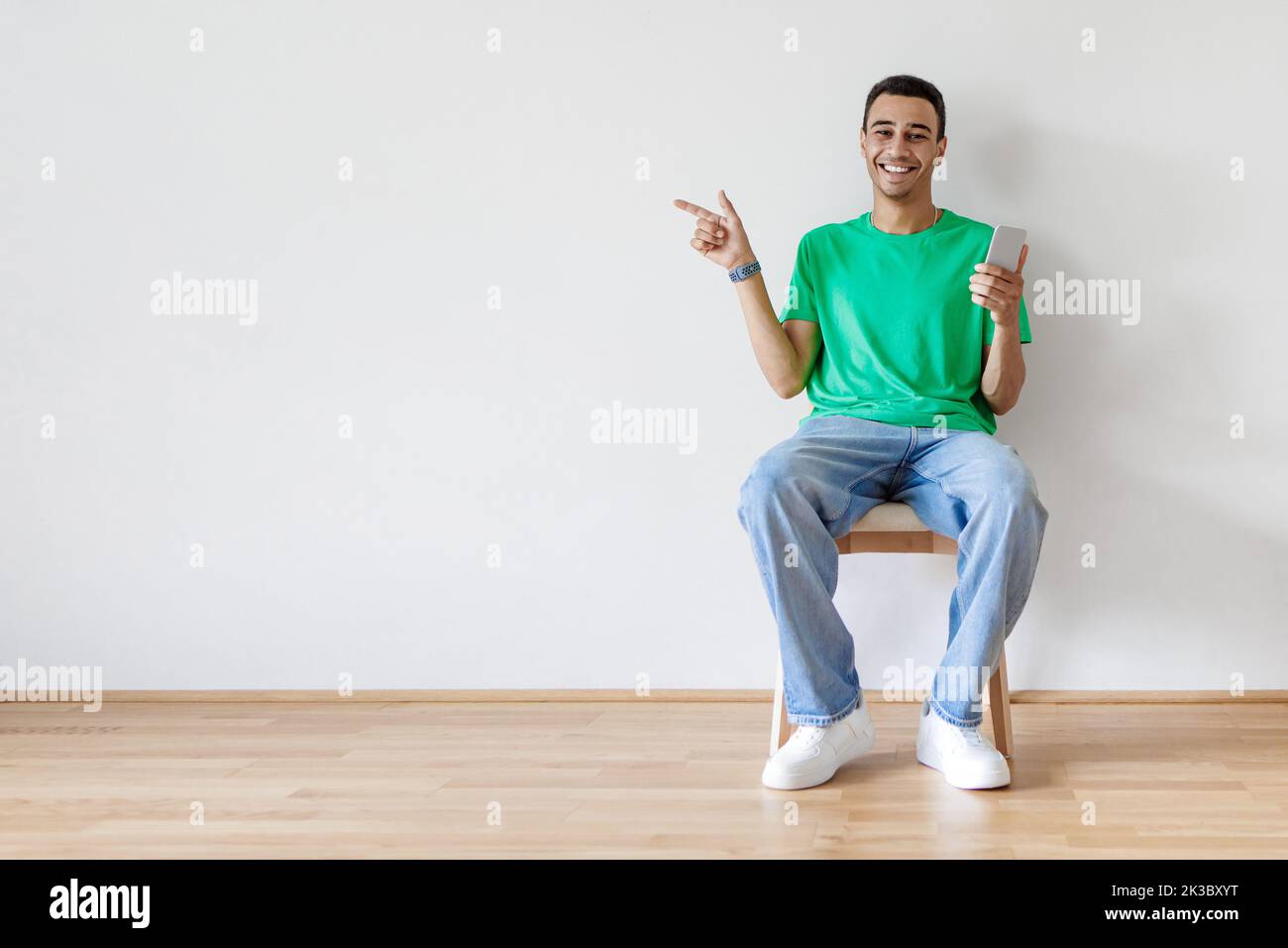 Felice giovane uomo arabo seduto sulla sedia con il nuovissimo smartphone e puntando da parte a copia spazio Foto Stock