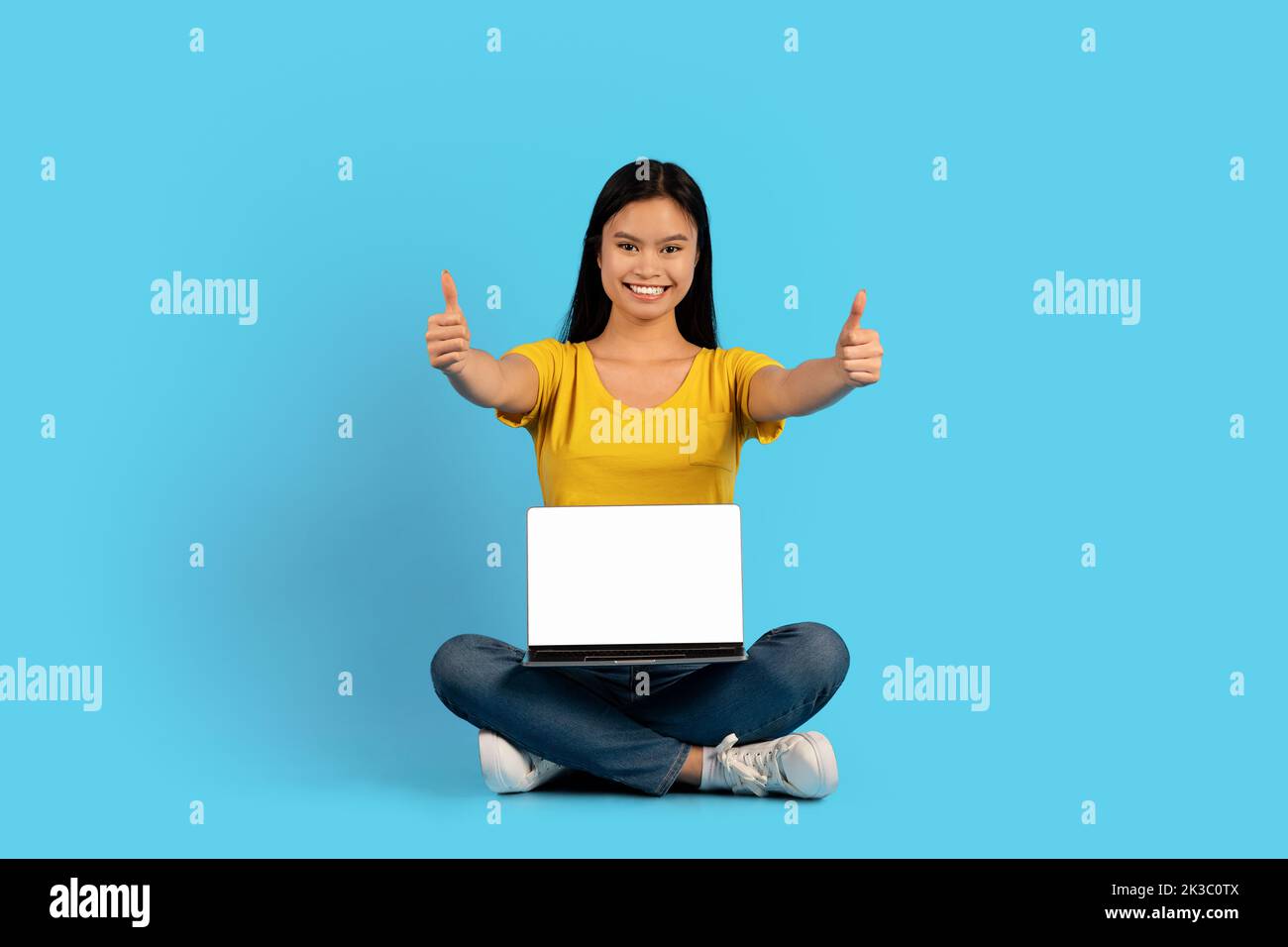 Felice giovane giapponese femmina siede in posizione lotus con computer portatile con schermo vuoto, mostra pollici in su Foto Stock