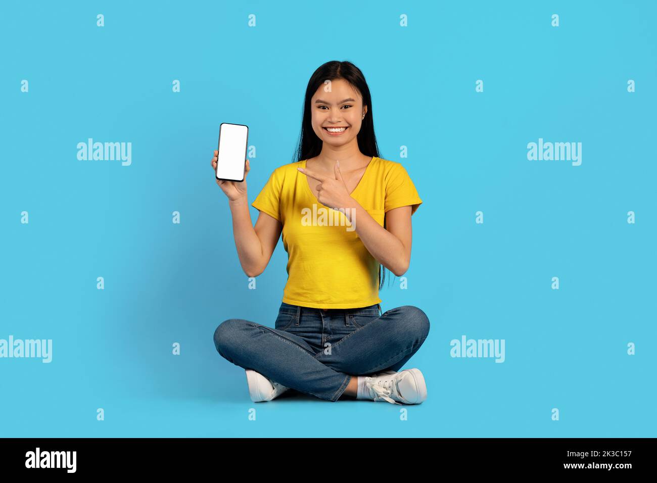 Giovane donna coreana sorridente in t-shirt gialla seduta in posizione lotus, puntando il dito verso lo smartphone con schermo vuoto Foto Stock