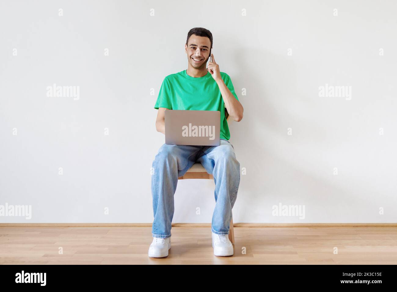 Ritratto completo di un ragazzo con un computer portatile che comunica sul telefono cellulare, seduto su una sedia contro una parete bianca, spazio copia Foto Stock