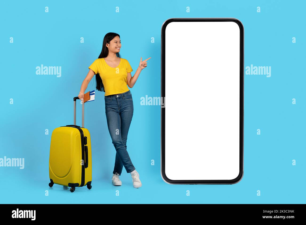 Giovane donna coreana sorridente in una t-shirt gialla con passaporto, biglietti aerei, punta il dito sull'enorme smartphone Foto Stock