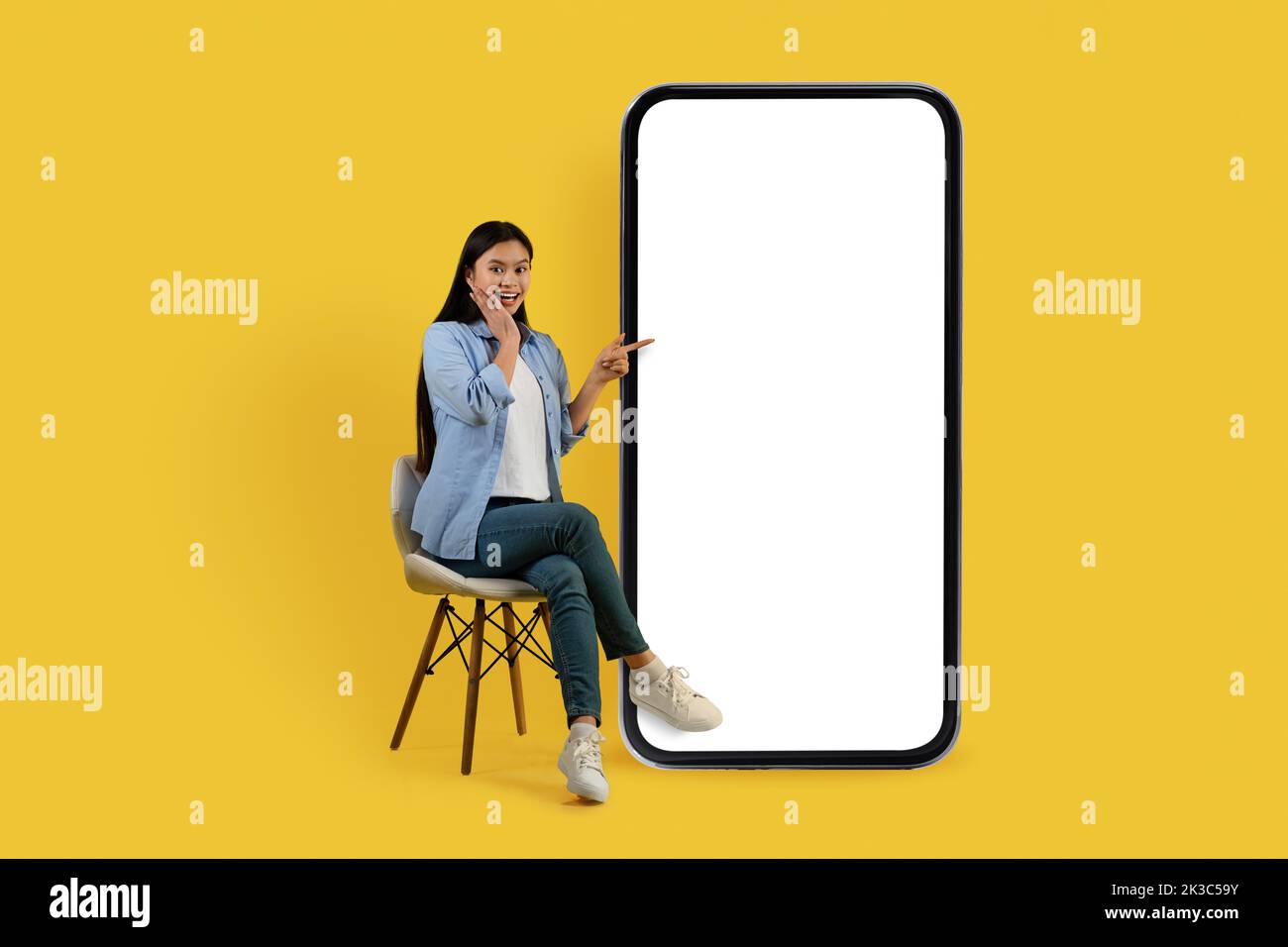 Sorridente scioccata giovane studentessa cinese in casual si siede sulla sedia, punta il dito contro enorme smartphone con schermo vuoto Foto Stock