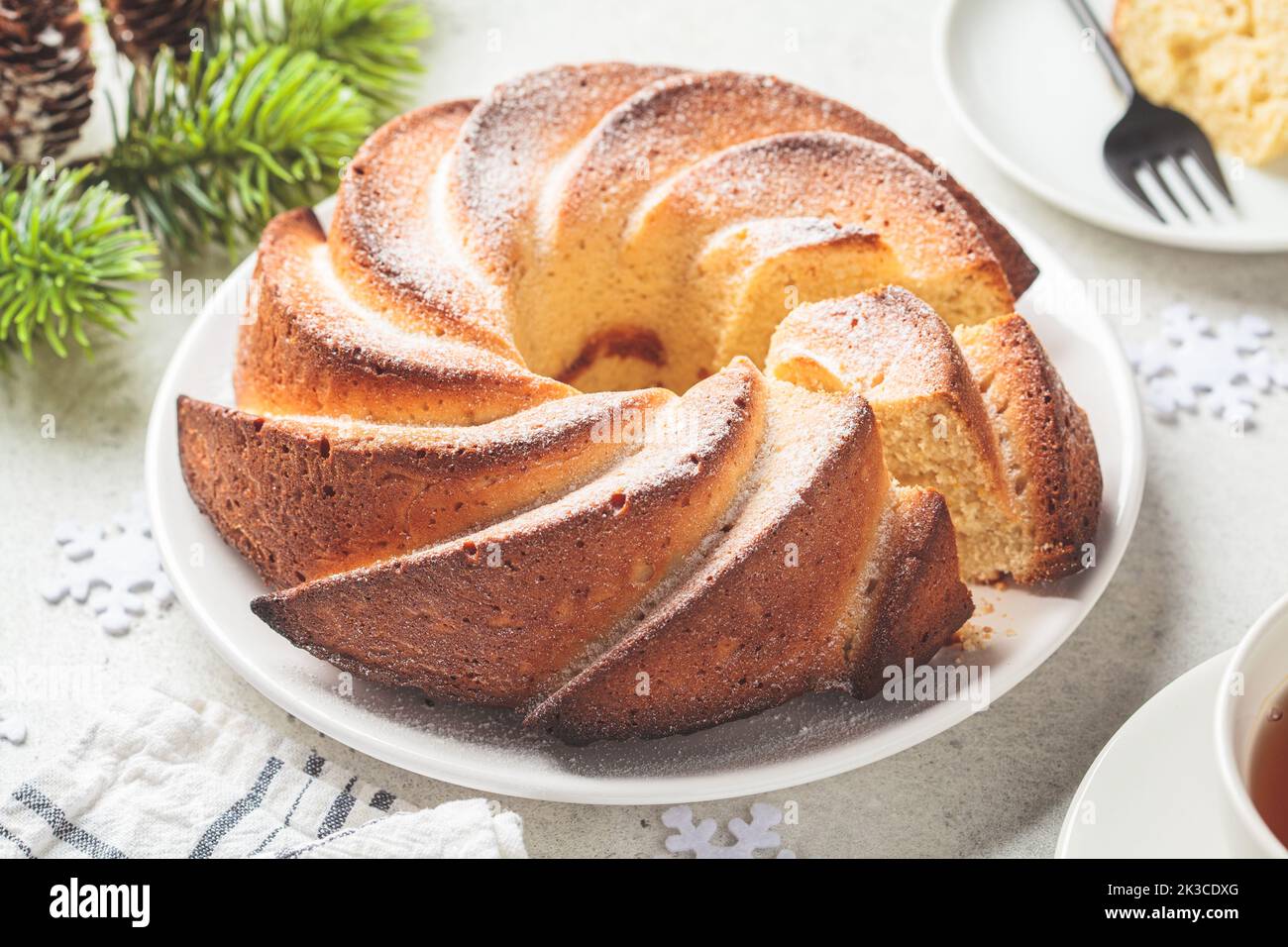 Dessert di Natale, primo piano. Torta alla vaniglia con zucchero a velo su un piatto bianco, fondo grigio chiaro con rami di albero di Natale. Foto Stock