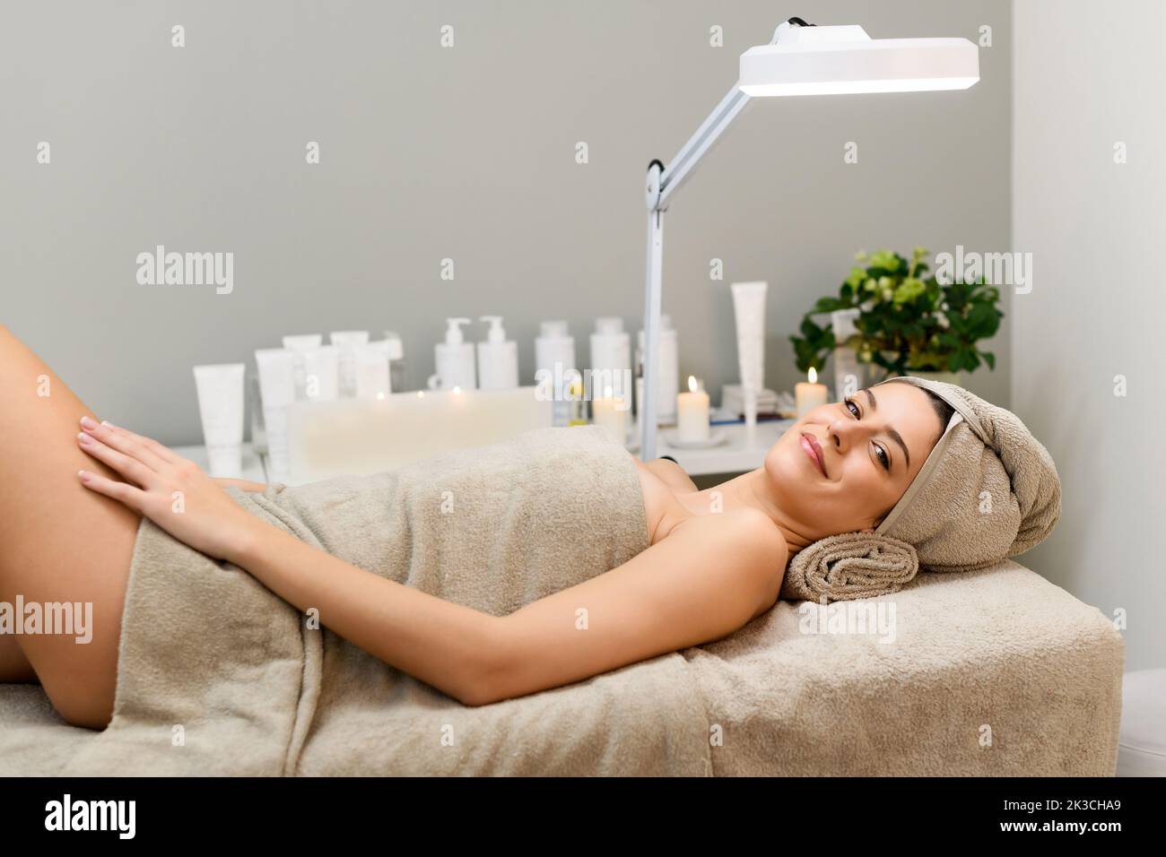 Angolo alto di felice cliente femminile coperto di asciugamano sorridente e guardando la macchina fotografica mentre sdraiato sul tavolo durante la sessione di bellezza nel salone termale Foto Stock