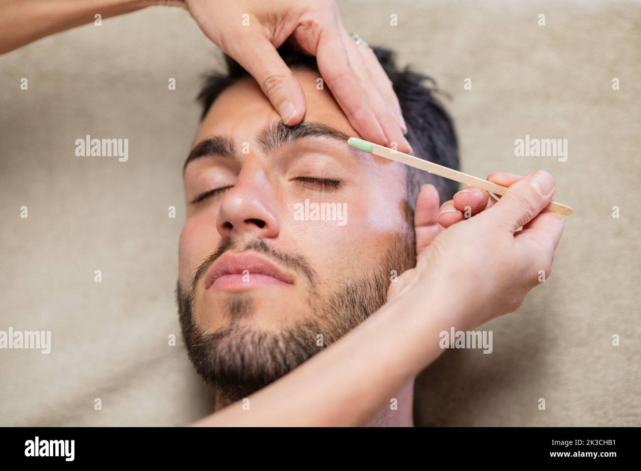 Dall'alto Crop donna usando bastone per applicare cera sul sopracciglio di giovane uomo bearded con gli occhi chiusi durante il lavoro in studio di bellezza Foto Stock