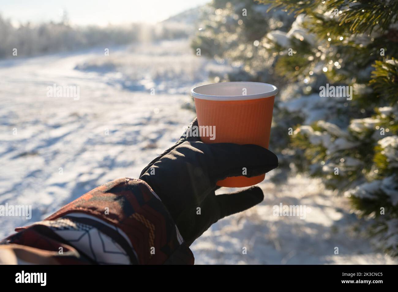 Tazza di tè viaggio invernale. Una tazza di carta arancione nelle mani dei guanti da donna. Inverno luminoso sfondo sfocato soleggiato. Il concetto di ricreazione attiva, viaggi, h Foto Stock