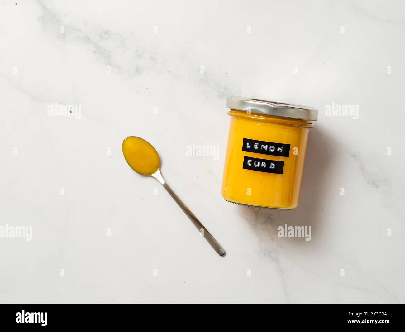 Deliziosa cagliata al limone fatta in casa. Vasetto di vetro con cagliata di limone e cucchiaio pieno su sfondo di marmo bianco. Etichetta adesiva nera con il testo Lemon Curd sul vaso. Vista dall'alto o disposizione piatta. Spazio di copia Foto Stock