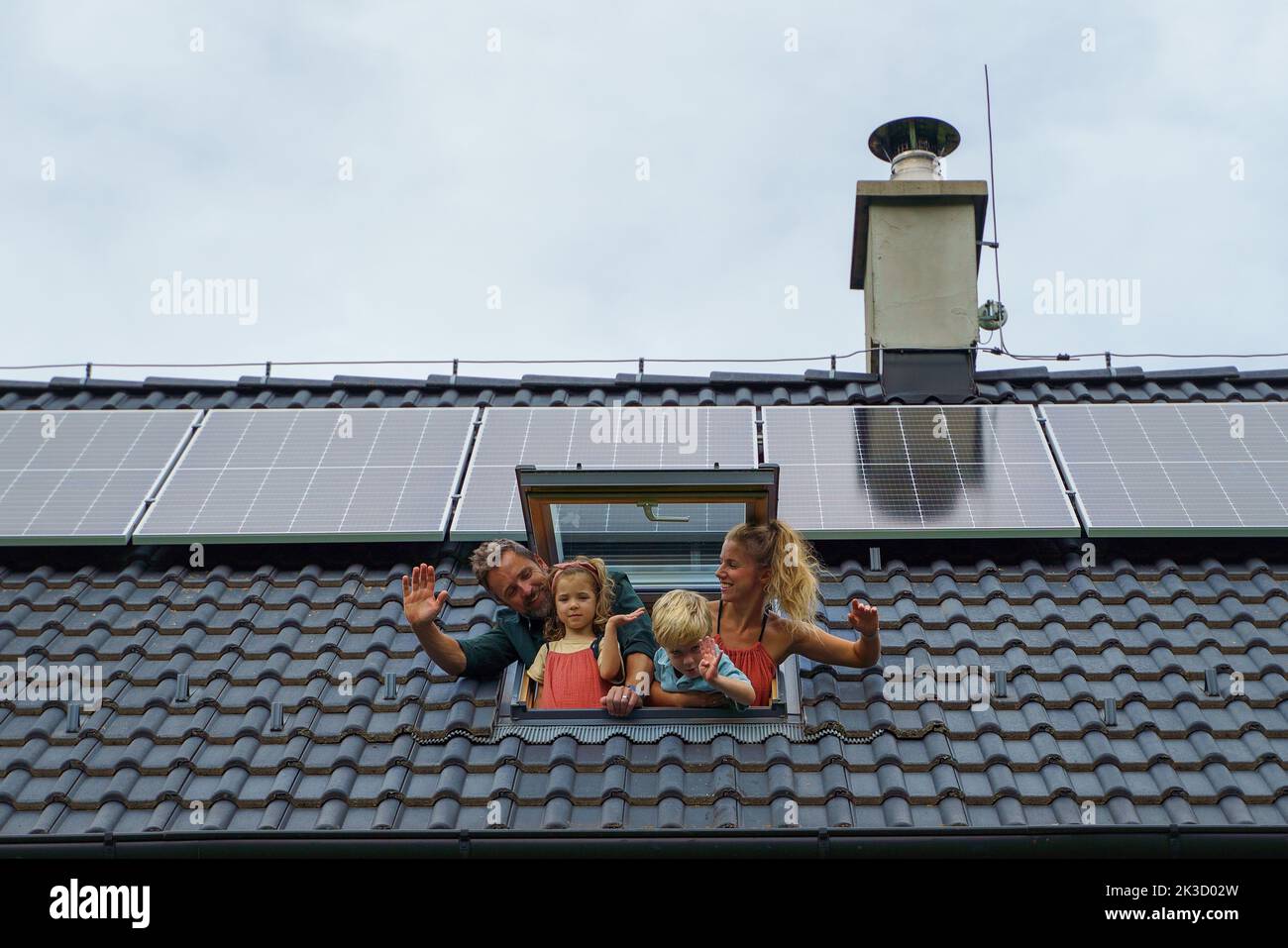 Felice famiglia che sventolava dalla finestra del lucernario nella loro nuova casa con pannelli solari sul tetto. Energia alternativa, risparmio di risorse e sostenibilità Foto Stock