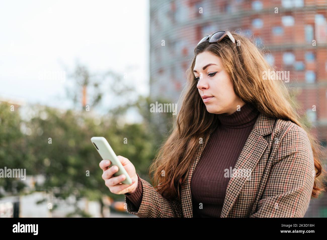 Donna in outerwear con capelli lunghi che navigano sui social media sul cellulare mentre si trova in piedi su sfondo sfocato della strada cittadina di giorno Foto Stock