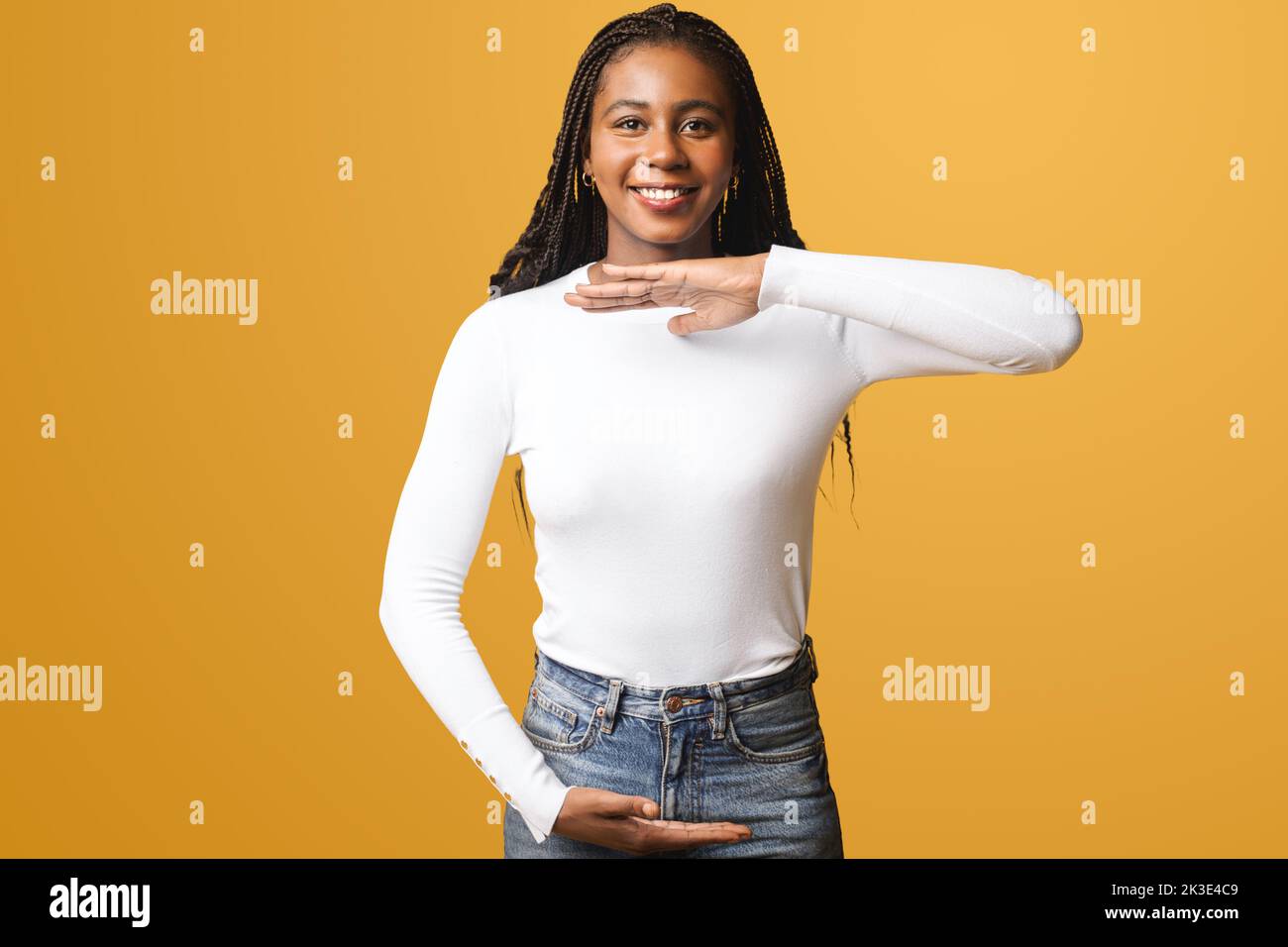 Sorridente giovane afroamericana con capelli afro indossare casual elegante su sfondo giallo tenendo vuoto con le mani, misurare simbolo, sorridere e guardare la macchina fotografica, pubblicità concetto Foto Stock
