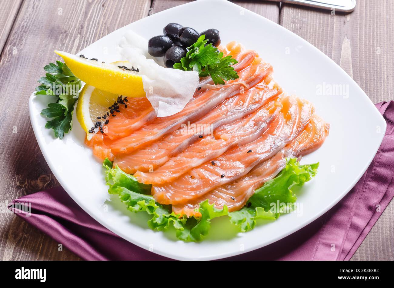 Pesce rosso fresco tagliato a strisce con limone ed erbe su un piatto bianco su uno sfondo di legno scuro Foto Stock