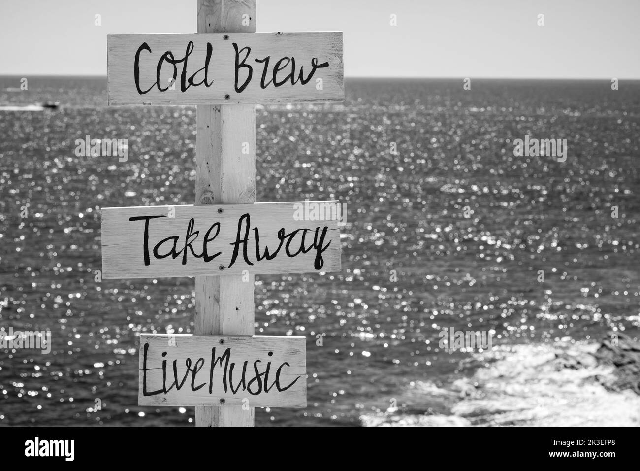 Vista di segni di legno per take away, Cold Brew, musica dal vivo e il mare sullo sfondo sulla spiaggia di Mylopotas in iOS Grecia in bianco e nero Foto Stock